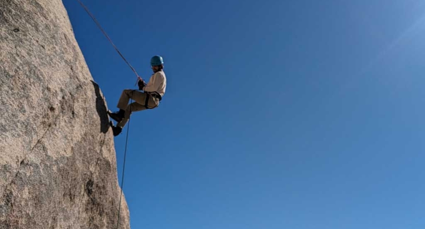 a person wearing safety gear is secured by ropes on a rock wall below a blue, cloudless sky. 